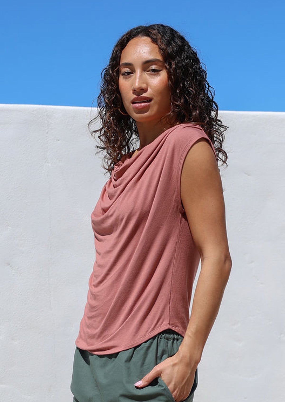 Woman wearing a pink sleeveless top with draped neck against a white wall with blue sky.