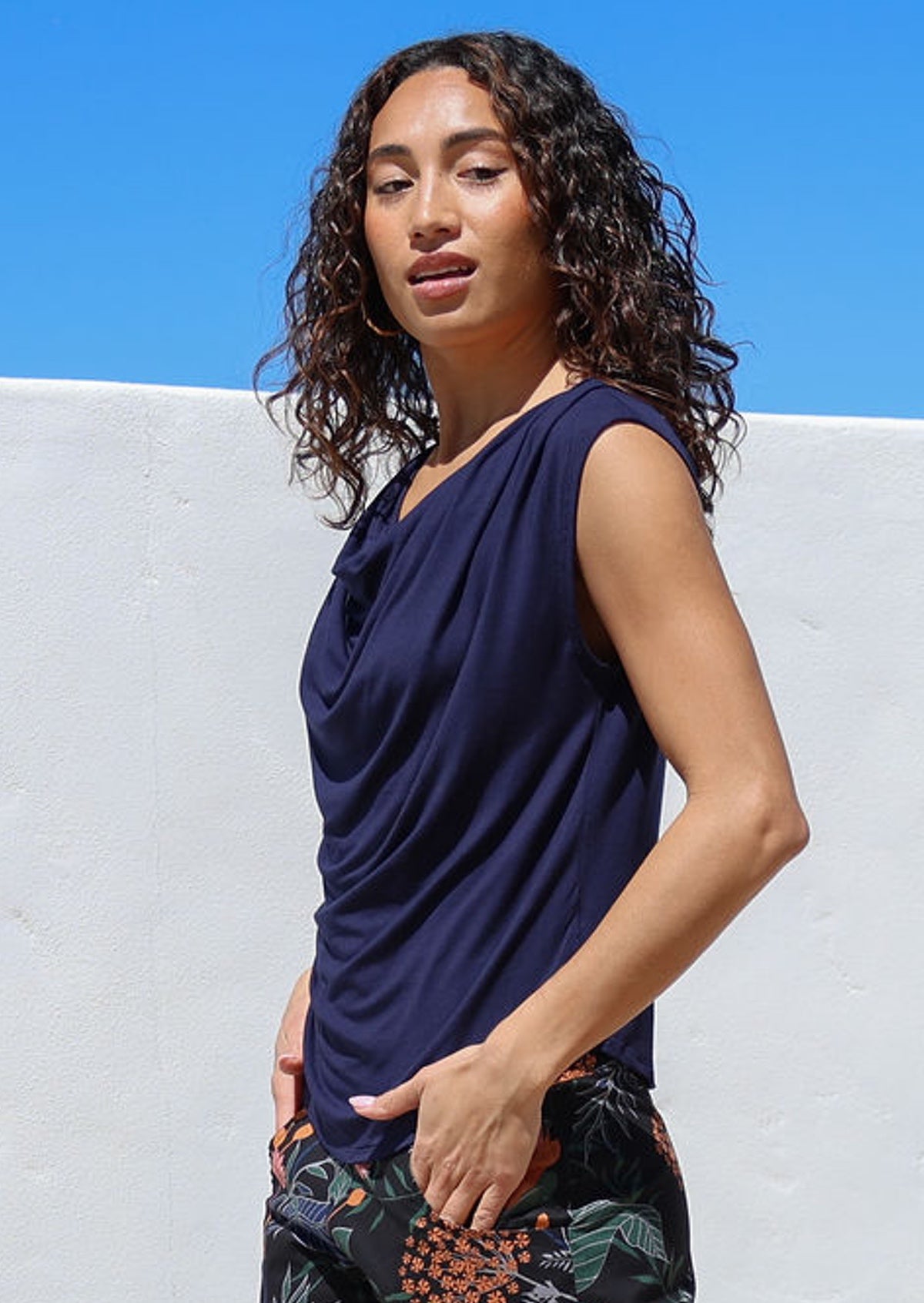 Woman wearing a navy blue sleeveless top with a cowl neckline, against a white wall with blue sky.