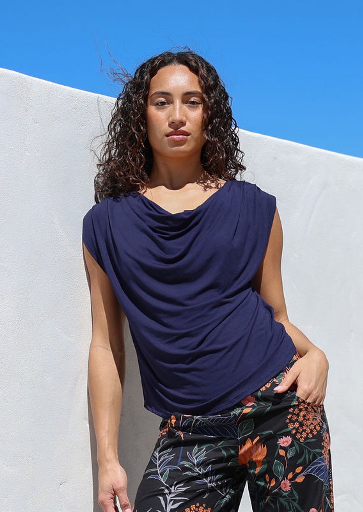 Woman wearing a blue sleeveless top and patterned pants against a white wall with hand in pocket of pants