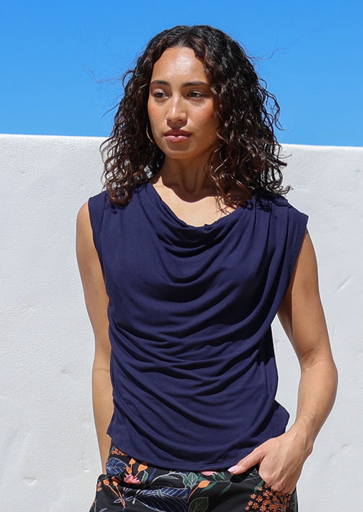 Woman wearing a navy blue sleeveless top with pleats on the shoulders and a cowl neckline