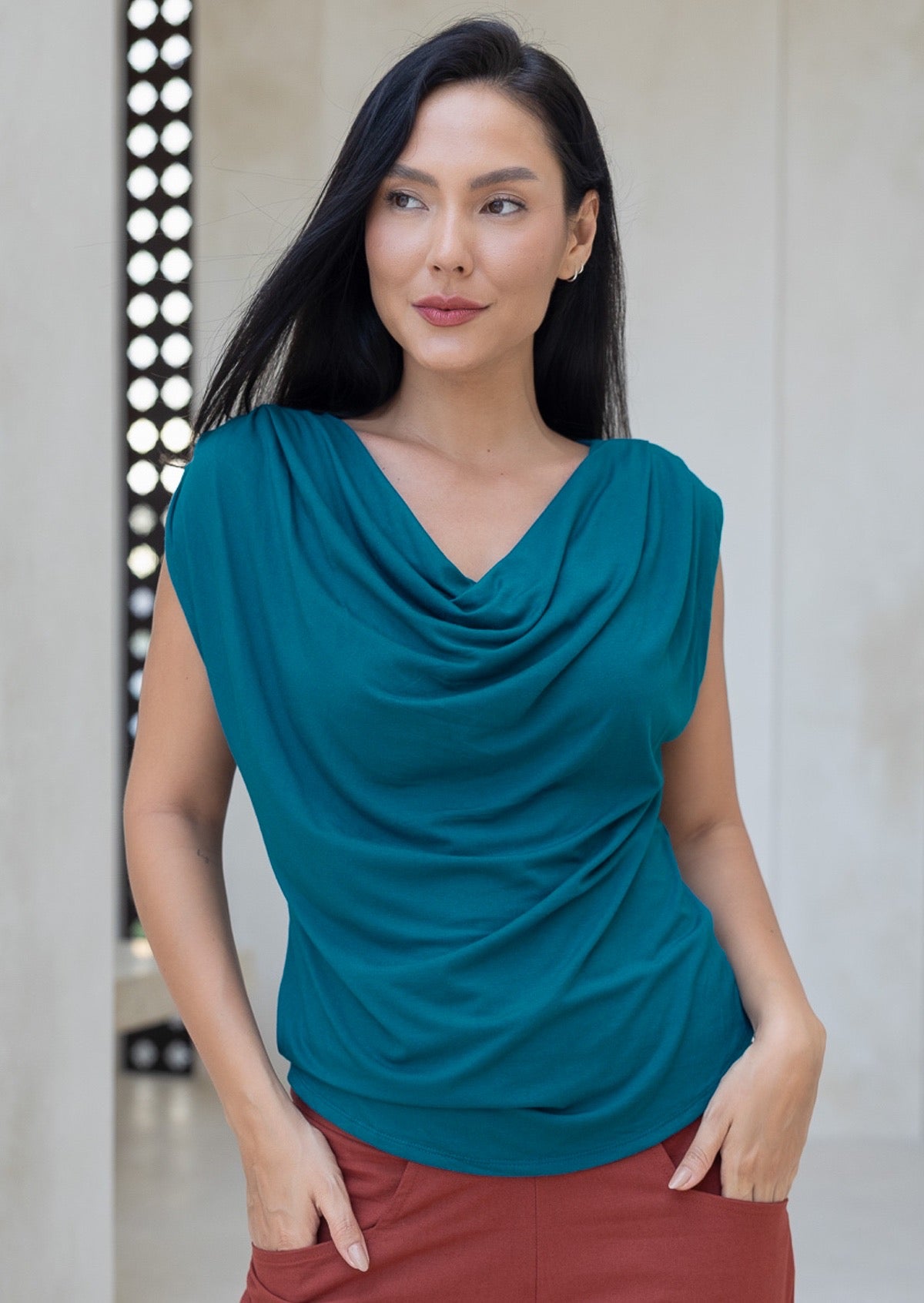 Woman wearing a teal blue top with a draped neckline, paired with rust pants with hands in pockets