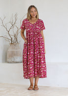 Woman wearing a pink floral  cotton dress standing against a white wall with a vase and branches in the background.