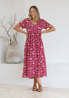 Woman wearing a 100 % cotton pink floral dress standing against a white wall with a vase and branches in the background.