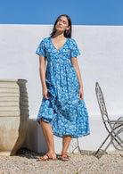 Woman wearing a blue floral dress cotton maxi dress standing against a white wall with a blue sky.
