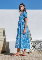 Woman wearing a cotton maxi dress blue floral cotton standing against a white wall with a clear blue sky.