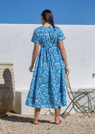 Woman wearing a blue floral maxi dress standing in front of a white wall with a blue sky.