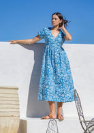 Woman in a blue cotton floral sundress standing against a white wall with a clear blue sky.