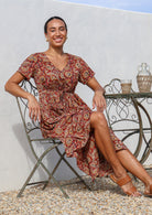 Model seated on a chair wearing a lightweight cotton relaxed fit button through floral dress with V neckline and a shoestring tie.