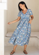 Woman wearing a blue based floral cotton midi length dress, with hand in pocket, swishing to one side