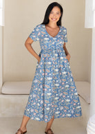 Woman wearing a blue based floral button through dress with a V-neckline and short sleeves, with hands in pockets