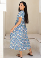 Back view of woman wearing a blue based floral print cotton midi length dress with thin side ties, tied at the back