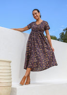 Woman standing with crossed legs in purple cotton, button through floral maxi dress with V neck, short sleeves and pockets.