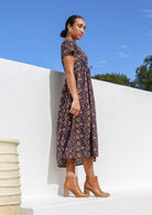 Model posing on side wearing a button through cotton dress with short sleeves and pink floral print on dark purple base.