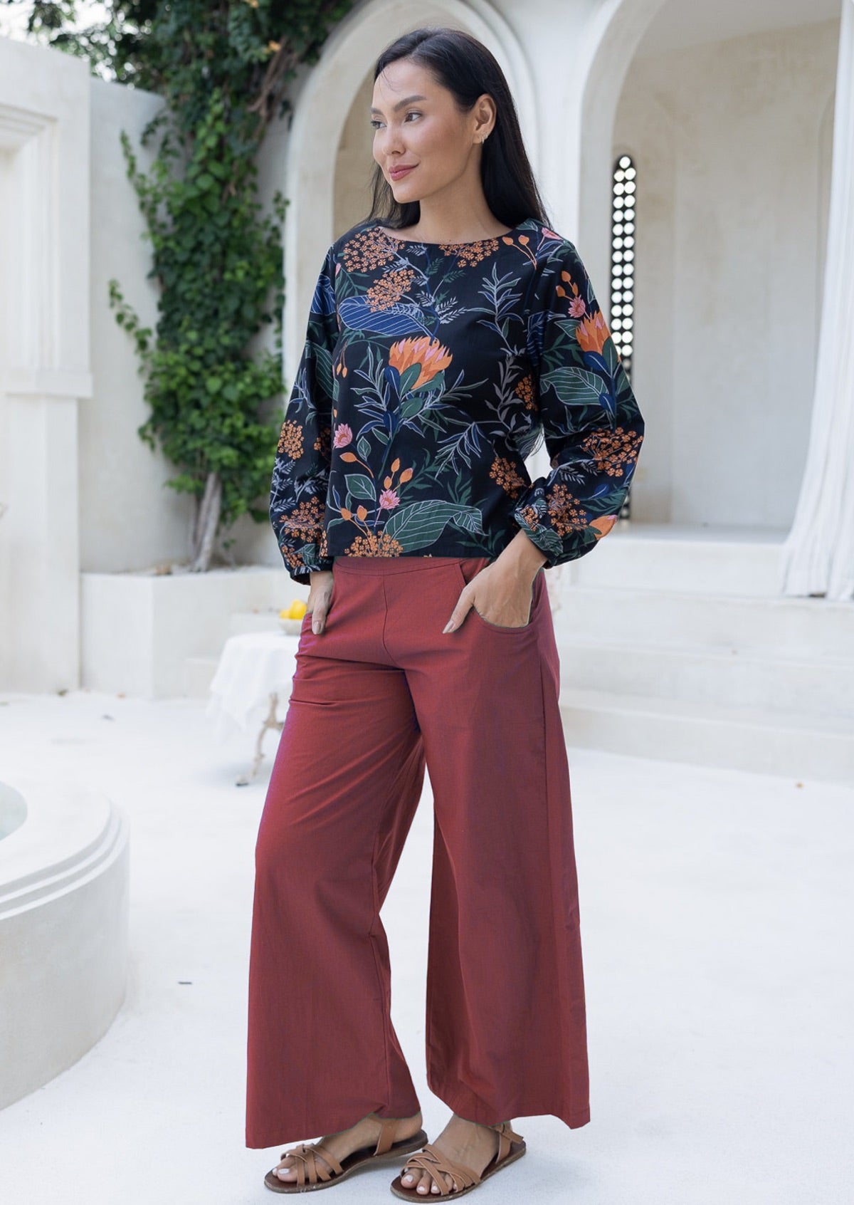 Woman wearing a floral top and rust colour cotton pants standing in a white architectural setting.