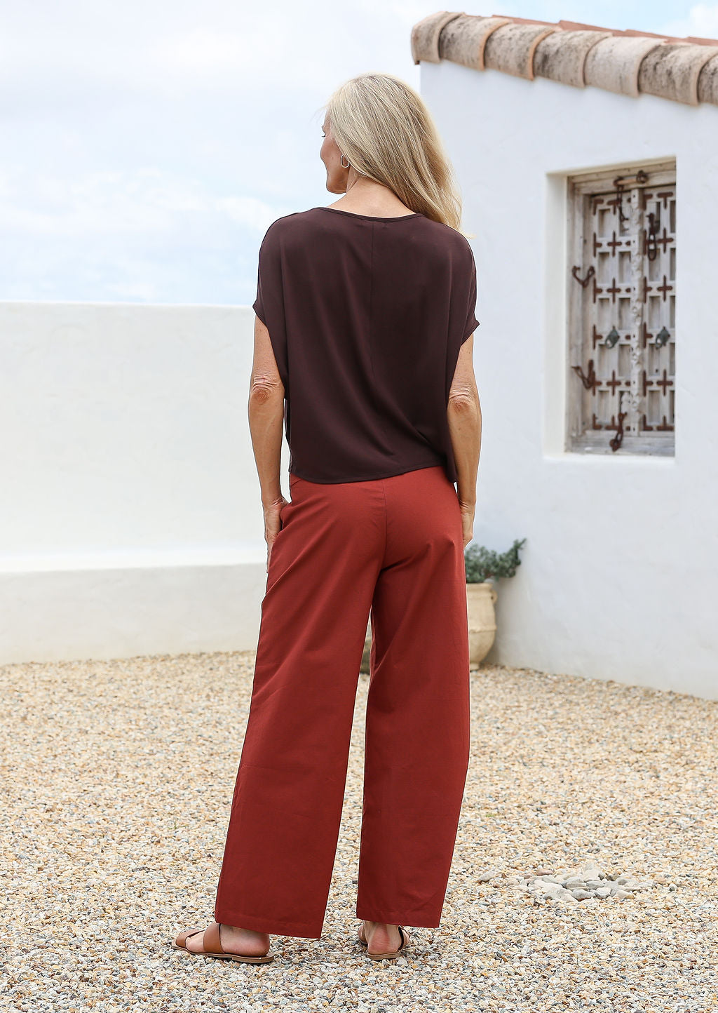 Woman standing in a back pose with hands in pocket wearing a relaxed fit 100% cotton pants in rust shade with side zipper and high waisted style