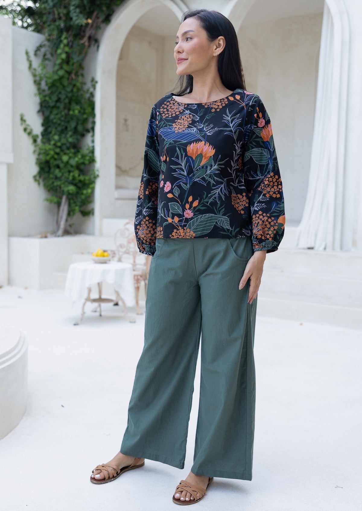 Woman wearing a floral patterned top and green wide leg pants standing in a white outdoor setting.