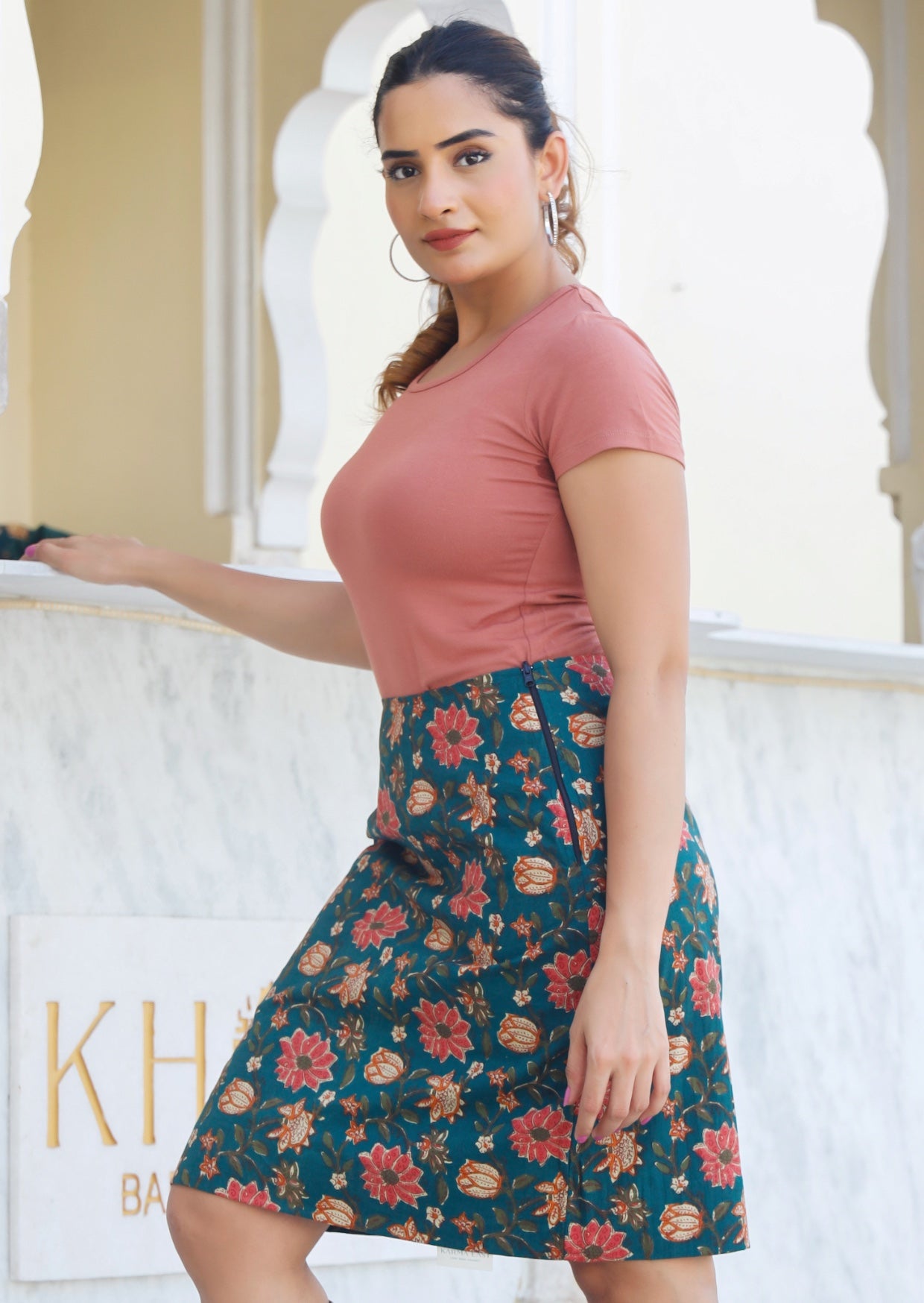Model standing sideways wears a reversible cotton skirt in bold floral print on green base with short sleeve basic top in dusty rose