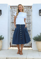 Woman standing in front of decorative wall wears 100% cotton knee length button through skirt in dark sea blue with box pleats and hands in pockets