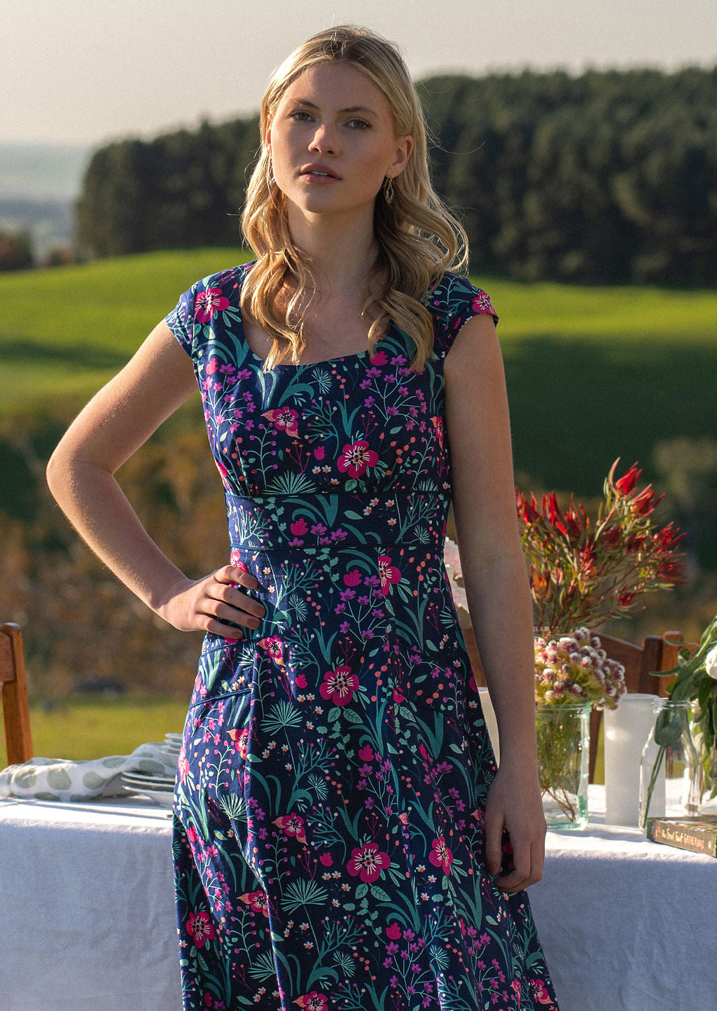 woman in retro inspired Scarlett Dress Dianthus in a  vibrant navy, purple, pink and aqua floral print on 100% cotton in front of laid out table