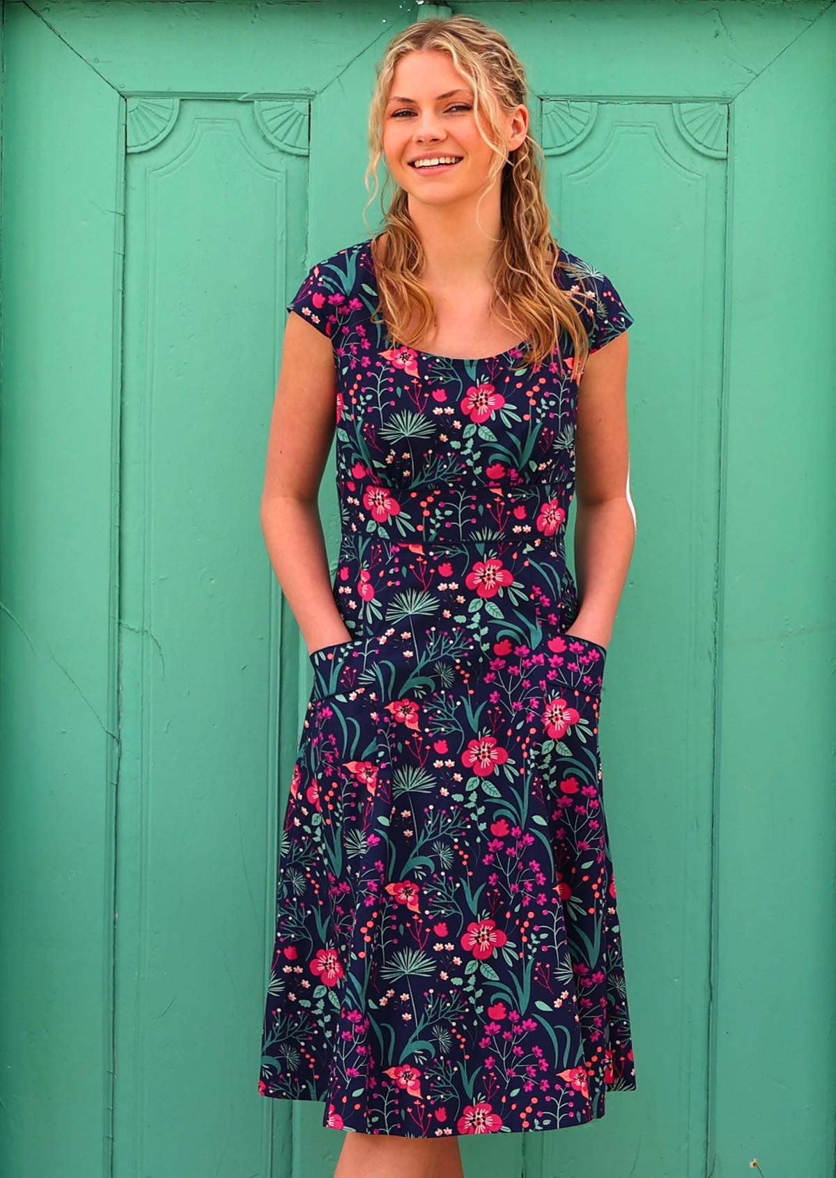 woman in front of aqua door in retro inspired Scarlett Dress Dianthus  vibrant navy, purple, pink and aqua floral print on 100% cotton