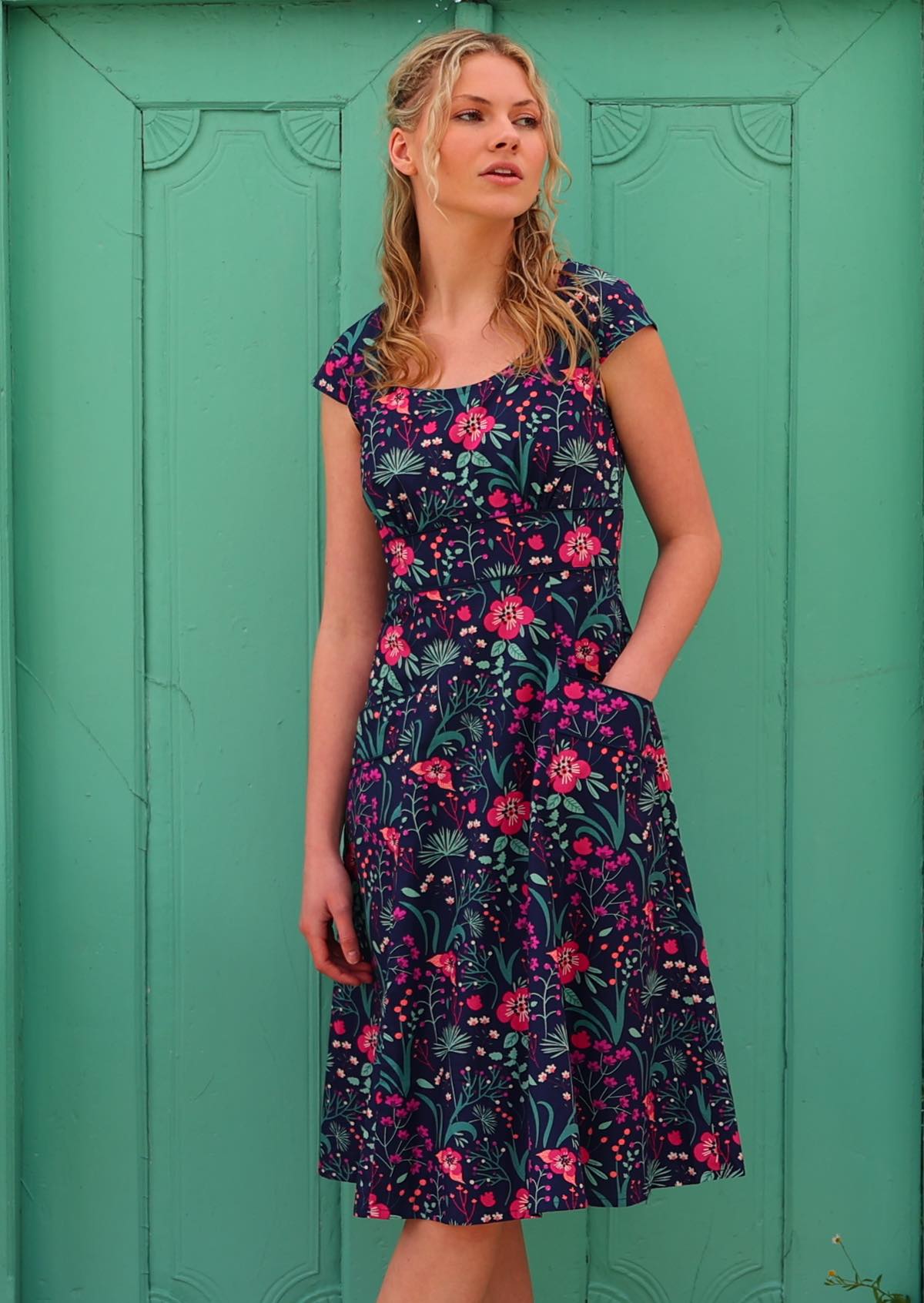 woman in retro inspired Scarlett Dress Dianthus in a  vibrant navy, purple, pink and aqua floral print on 100% cotton with hand in pocket in front aqua door