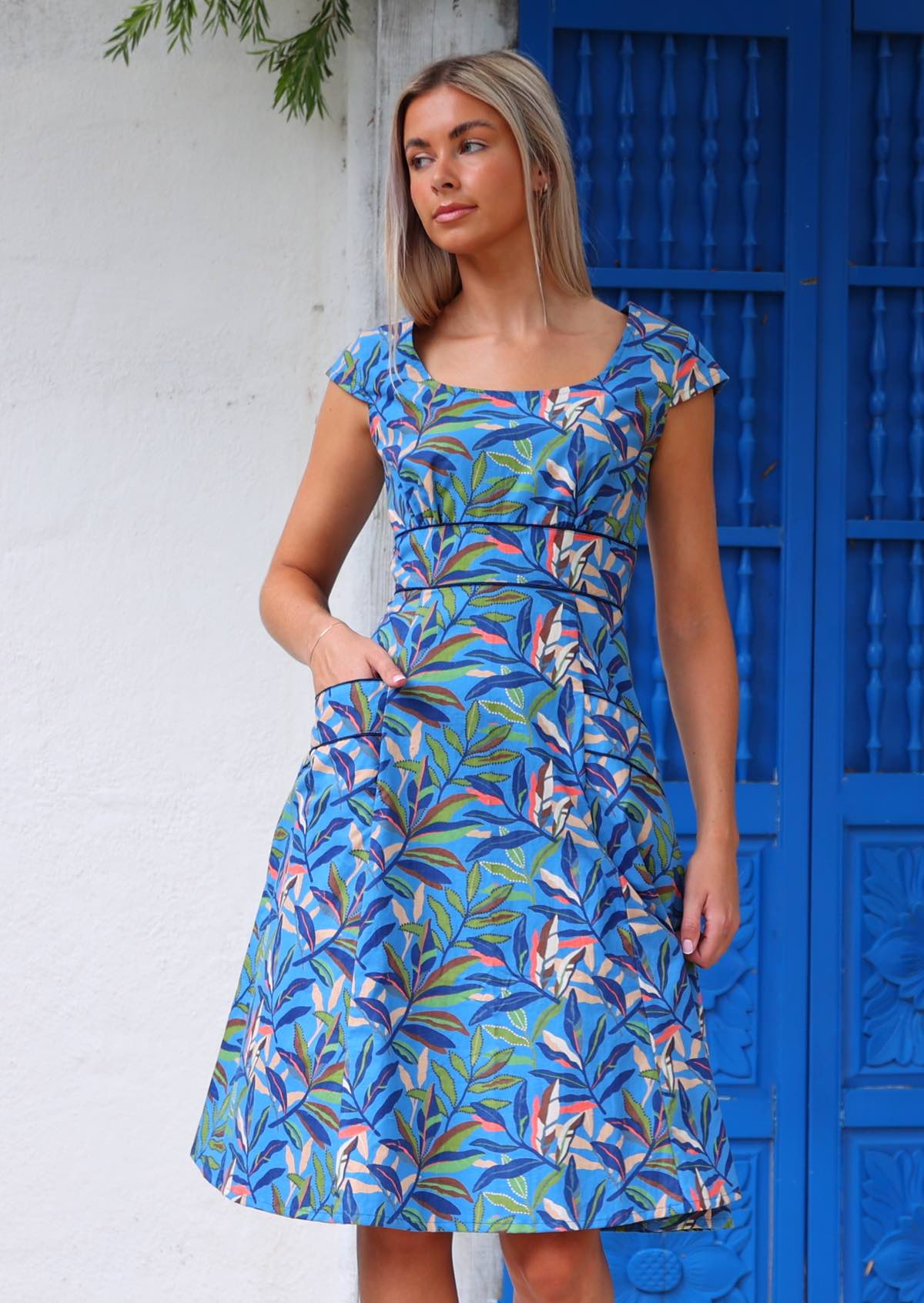 Woman wearing blue cotton retro style dress with pockets in front of blue door with hand in pocket