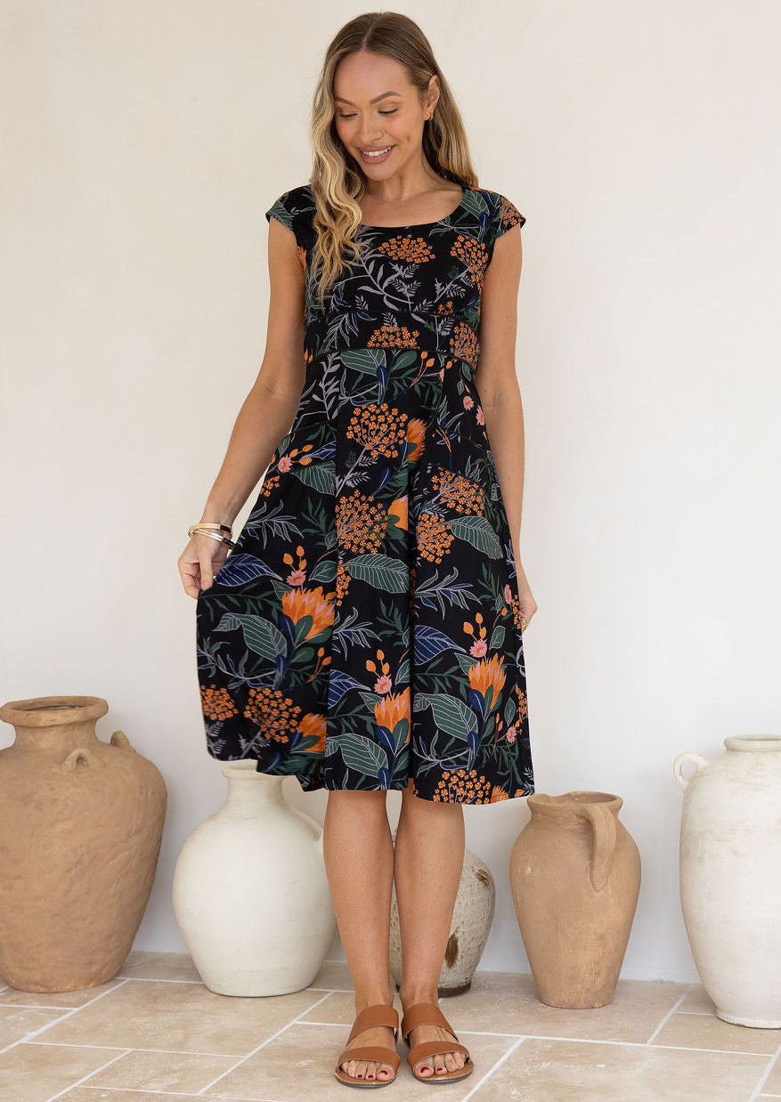 Model wearing a botanical print dress with scoop neck, cap sleeves and piped detailing at the waist and pockets.