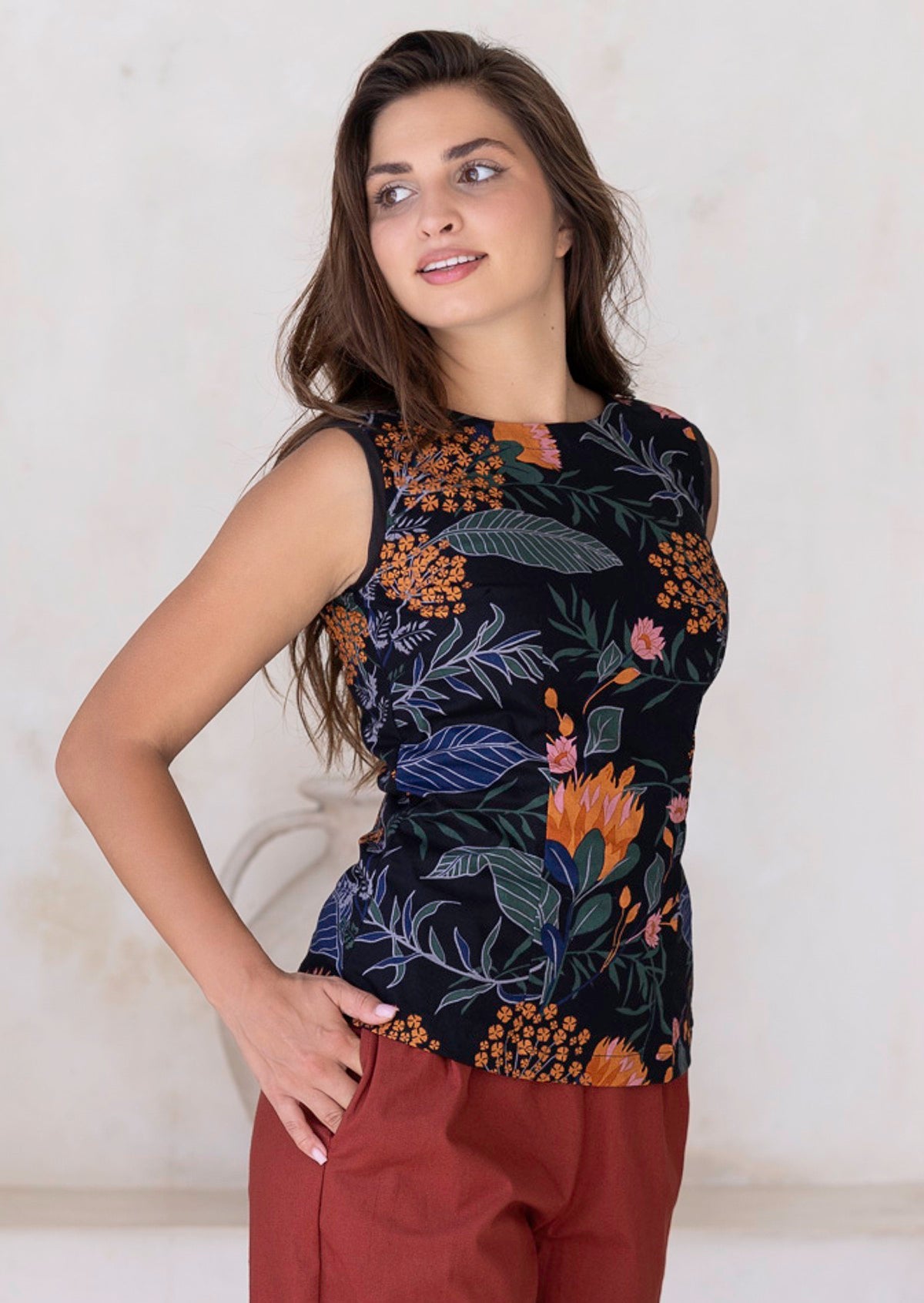 Woman wearing a sleeveless top with floral pattern against a plain background with her hand in pants pocket