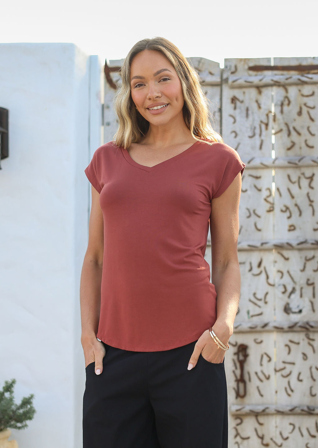 Woman wearing a 100% rayon terracotta V-neck T-shirt and black pants, standing against a white wall.