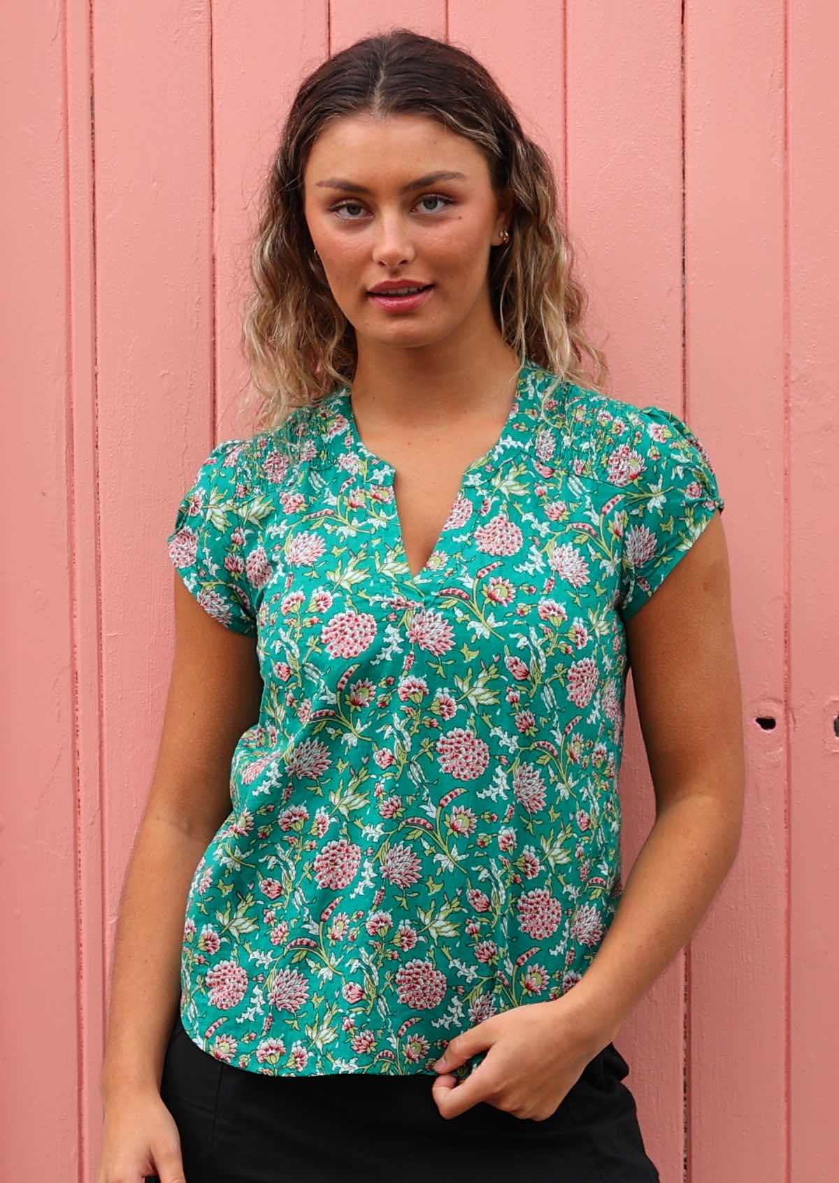Woman stands in cotton floral print cap sleeve top with V-neckline and shirring across the front of the shoulders