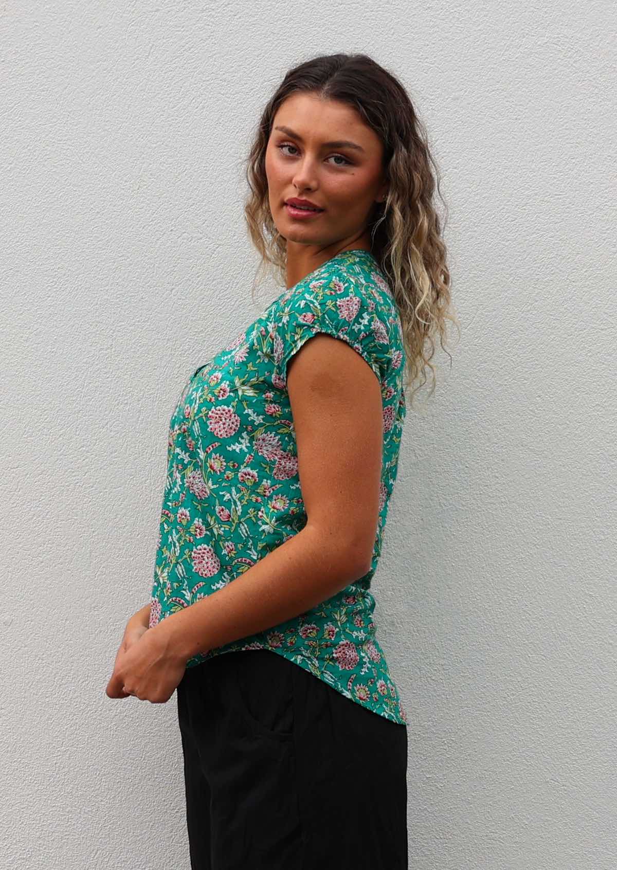 Model stands sideways in 100% cotton cap sleeve jade green based floral print top that is longer at the back