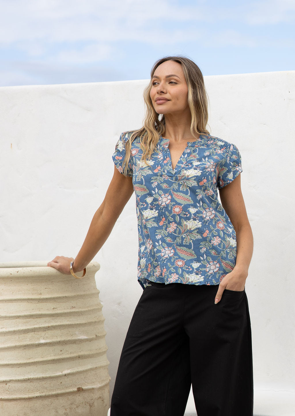 Woman posing in a dusty blue floral 100% cotton blouse with black cotton pants, standing against a white wall.