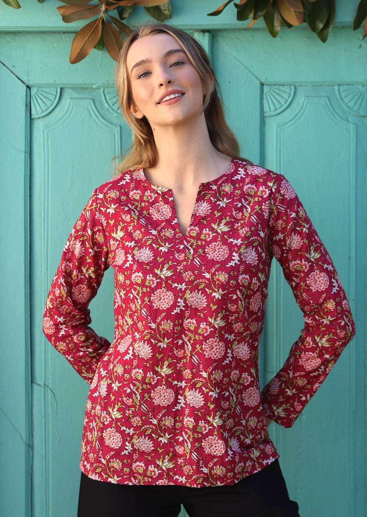 Model standing in front of aqua door wears long sleeve cotton shirt top with round neckline and v cutout.