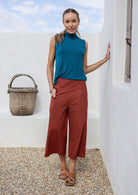 Woman in teal sleeveless top and rust 3/4 length pants standing against a white wall with a hand in pocket