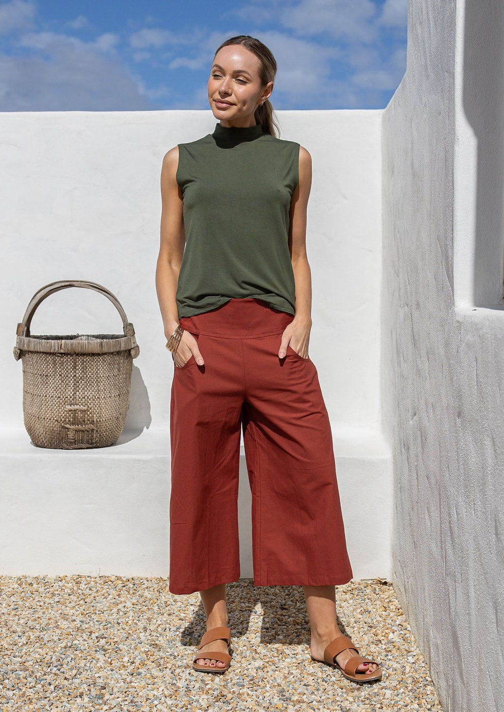 Woman wearing a green sleeveless top and rust 3/4 length pants standing against a white wall with hands in pockets
