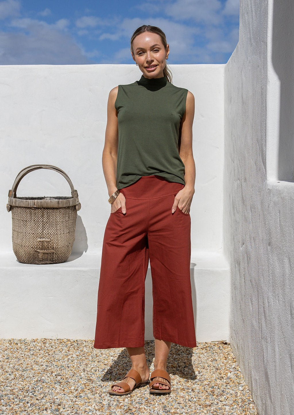 Woman wearing a green sleeveless top and rust 3/4 length pants standing against a white wall with a woven basket.