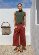Woman wearing a green sleeveless top and rust 3/4 length pants standing against a white wall with a woven basket.