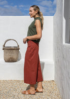 Woman in green top and rust 3/4 length pants standing against a white wall and blue sky