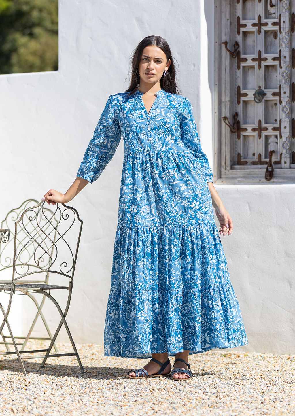 Woman wearing a blue cotton floral maxi dress with long sleeves standing outdoors against a white wall.