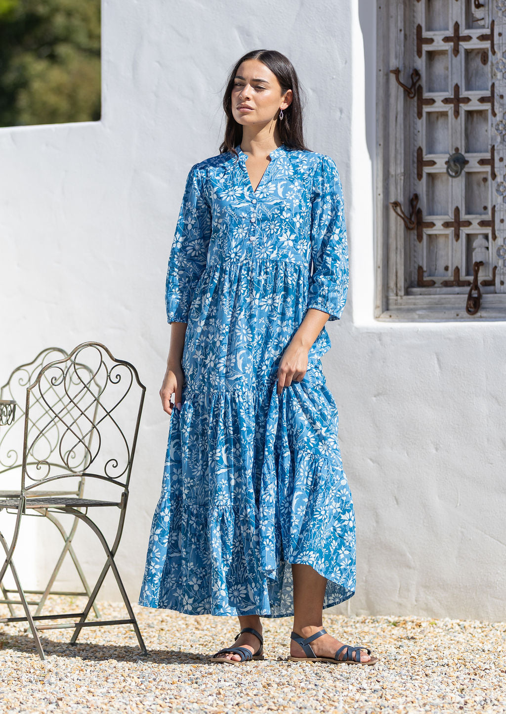 Woman wearing a blue cotton maxi dress with sleeves standing in front of a white wall with a metal chair.
