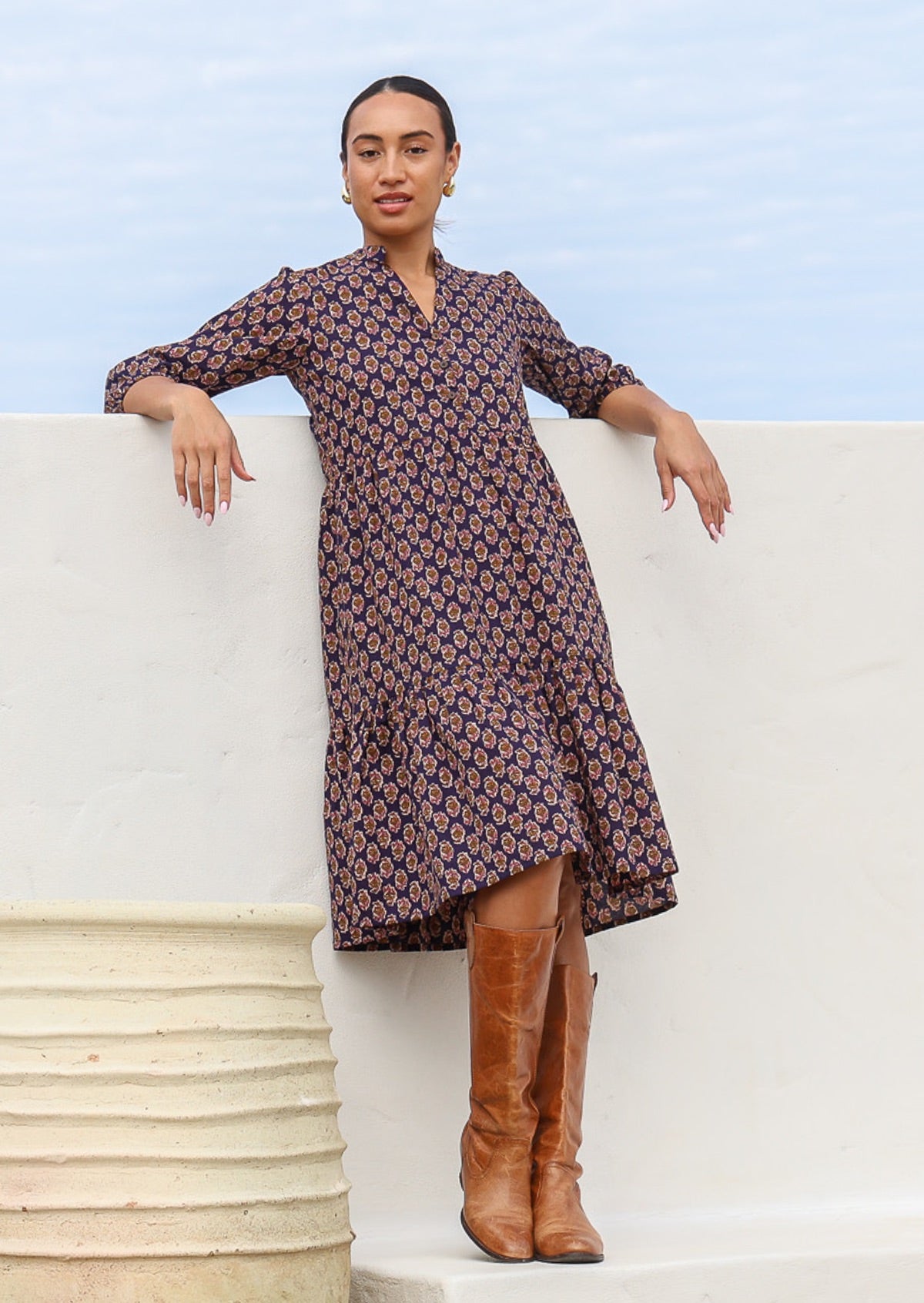 Woman leans on wall in cotton tiered over the knee length relaxed fit dress with 3/4 sleeves in a floral motif on a dark purple base