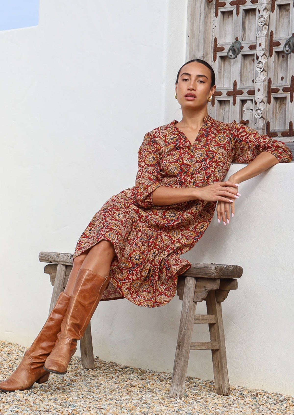 Woman sits on bench in cotton tiered dress in bright orangey red based floral print, with 3/4 sleeves, buttoned bodice and V-neckline
