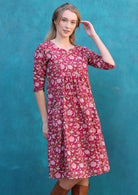 Model is wearing a loose fitted red cotton dress sitting just below the knees with empire waist and 3/4 sleeves.
