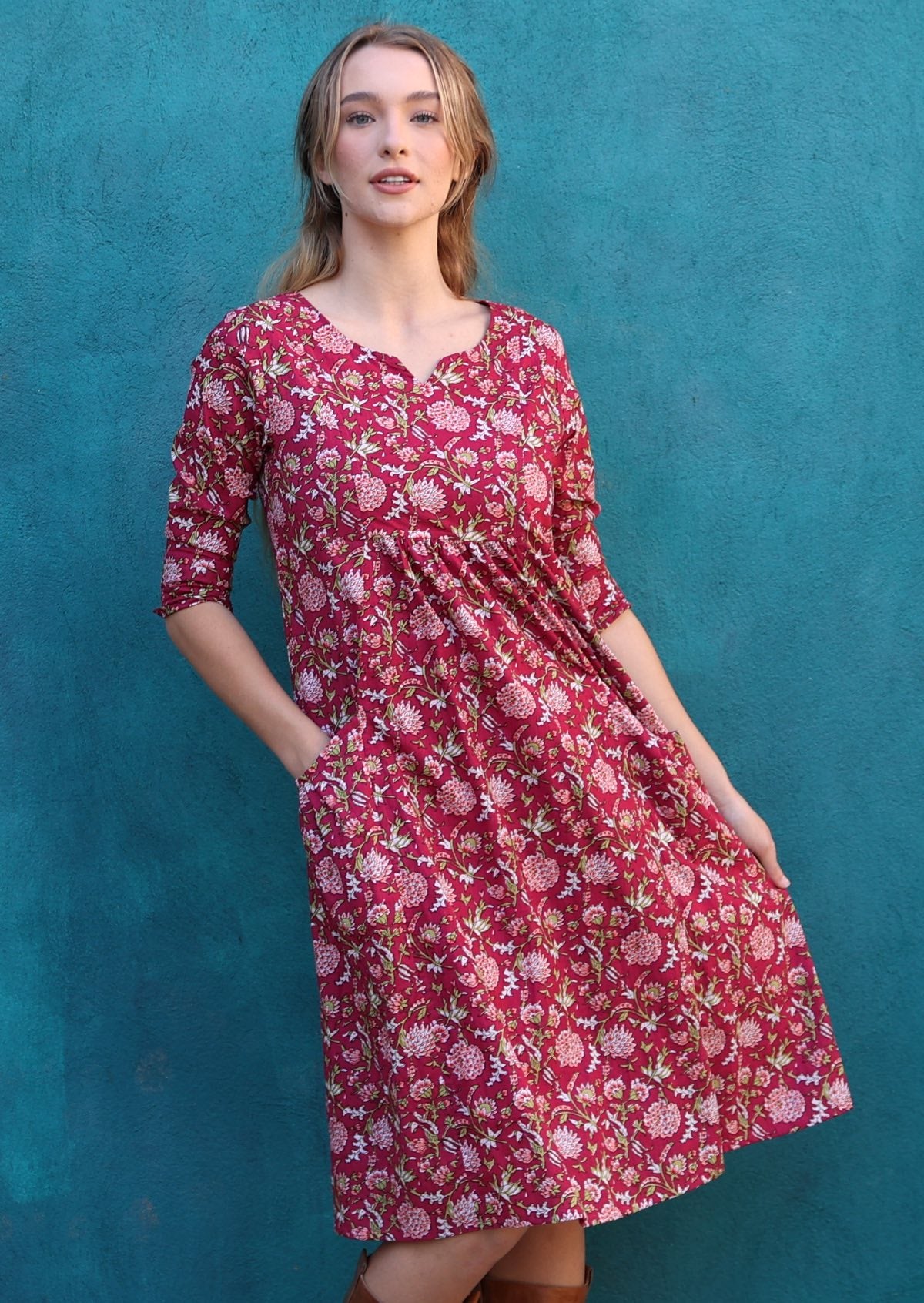 Model standing in front of blue wall wears  3/4 sleeves cotton dress with round neckline, pockets and pin tucks on waist.