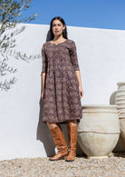 Woman wearing a brown cotton patterned dress and brown knee-high boots standing against a white wall with an olive tree.