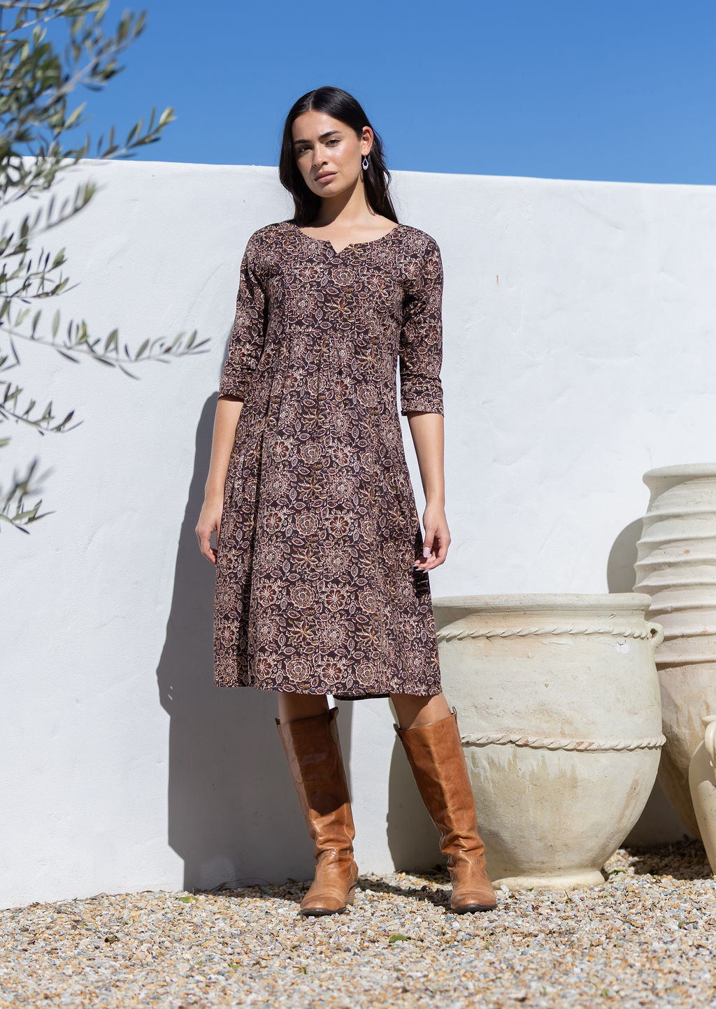 Woman wearing a brown cotton floral dress, with sleeves and brown boots standing against a white wall with greenery.