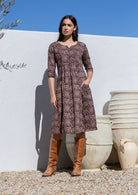 Woman wearing a brown floral cotton dress and brown boots standing against a white wall with large pots.