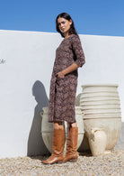Woman in a brown floral cotton patterned dress and brown boots standing against a white wall with hands in pockets
