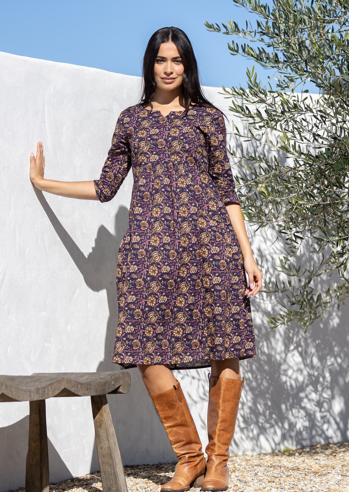 Woman wearing a purple cotton floral dress with brown boots standing outdoors against a white wall.
