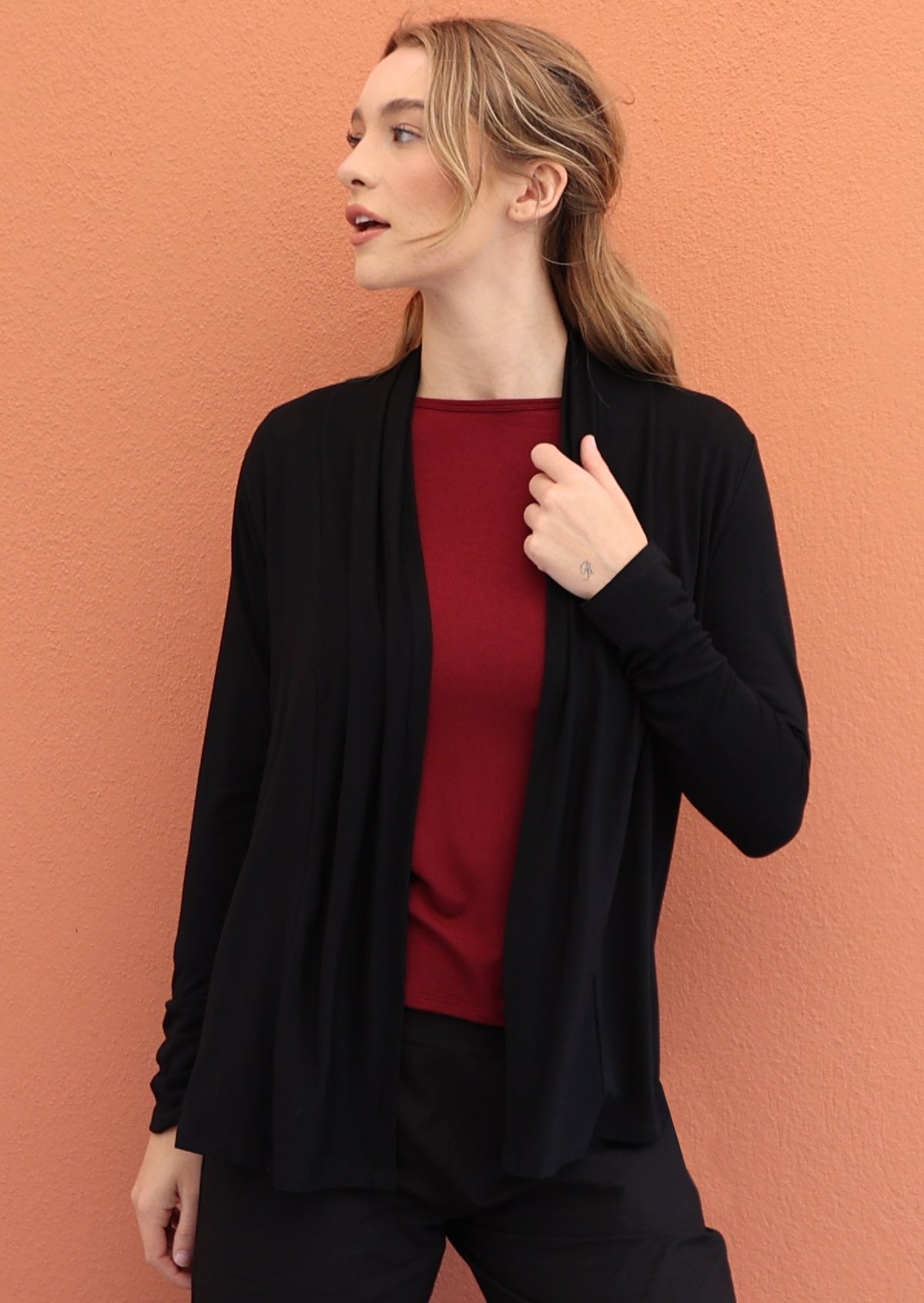 Woman wears black soft, stretch rayon cardi with long sleeves, holding the front opening revealing maroon top underneath
